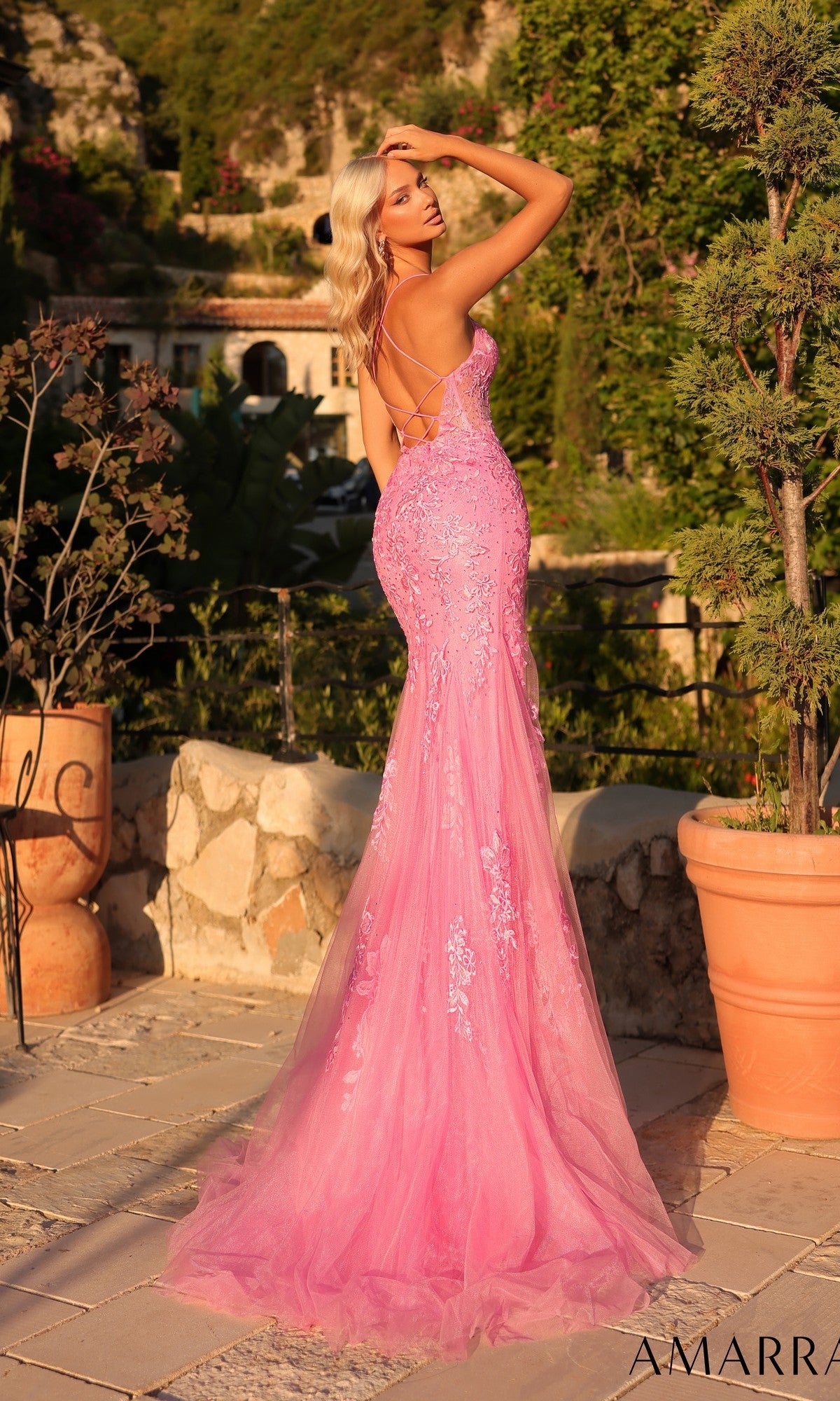 A blonde woman poses outdoors in the Amarra 89022 Embroidered-Lace Long Prom Dress, a fitted light pink gown with a lace-up back, surrounded by greenery and stone planters.